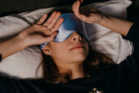 Woman lying on her back in bed wearing a blue silk sleep mask, resting her head on a white pillow.